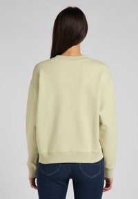 Sudadera de color verde claro con cuello ancho y puños acanalados, presenta un ajuste relajado y textura suave. Combina con vaqueros azul oscuro.