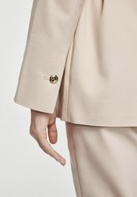 Beżowa marynarka o dopasowanym kroju, charakteryzująca się gładkim materiałem, eleganckim fasonem oraz złotym akcentem w postaci guzika. Ręka jest umieszczona w pobliżu rękawa marynarki.