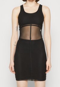 Žena v těsně padnoucích černých šatech bez rukávů se průhlednou středovou částí a viditelnými tetováními na levém paži.