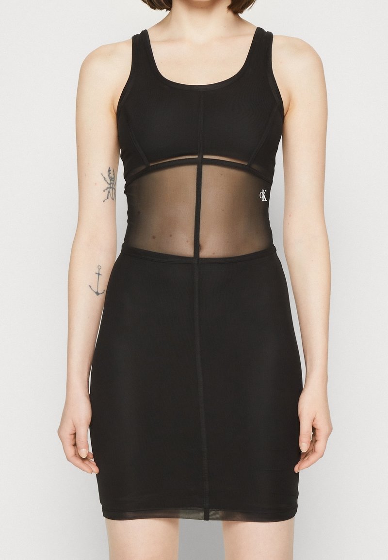 Žena v těsně padnoucích černých šatech bez rukávů se průhlednou středovou částí a viditelnými tetováními na levém paži.