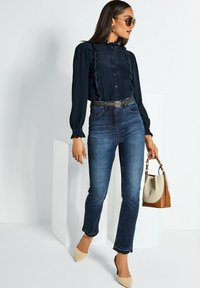 Frau in einer navyblauen Rüschenshirt, blauen Jeans, beigen Heels, Sonnenbrille und mit einer Tasche in Beige und Creme.