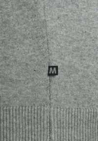 Pilkos megztinis su raukšlėta apatine dalimi. Pasižymi mažu juodu etiketu, ant kurio užrašytas baltas 'M' logotipas, pritvirtintas prie siūlės. Minkšta tekstūra.