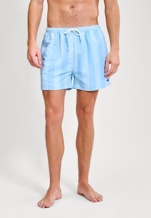 Hombre con bañador de rayas azules claras y cordón blanco, de pie descalzo contra un fondo liso y claro.