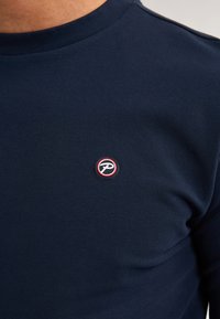 Navyblauwe katoenen t-shirt met een geribde ronde hals. Voorzien van een ronde logopatch met een rode rand op de linkerborst. Soepele textuur.