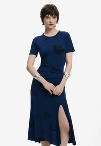 Mörkblå klänning med en åtsittande överdel, utsvängd kjol med en hög slits till låret och subtila svarta mönster. Mjuk, elastisk tyg.