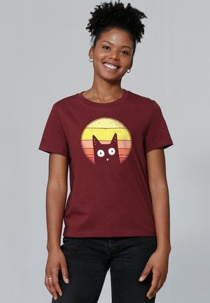 T-shirt en coton bordeaux avec un graphique de chat noir aux yeux blancs, sur fond d'un dégradé rétro circulaire de rayures jaunes et oranges.