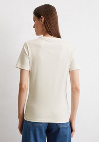Cremefarbenes T-Shirt mit kurzen Ärmeln und Rundhalsausschnitt, mit dezent gesticktem Logo auf dem Rücken, kombiniert mit blauen Jeans.
