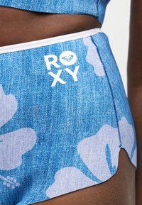 Short bleu à motif floral avec logo blanc "ROXY" en haut à gauche, porté par une personne à la peau foncée.