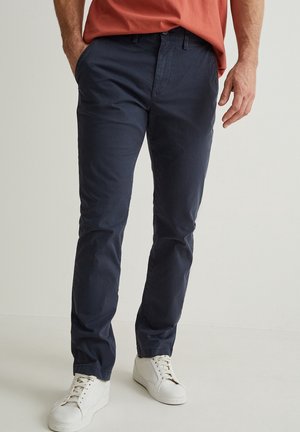 Uomo che indossa chinos blu navy, una t-shirt color ruggine e sneakers bianche, in piedi con una mano in tasca contro uno sfondo semplice.