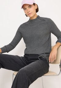 Grijze geribbelde gebreide sweater met hoge hals en zijkoorddetail, gecombineerd met zwarte broek. Model draagt een lichtroze pet.