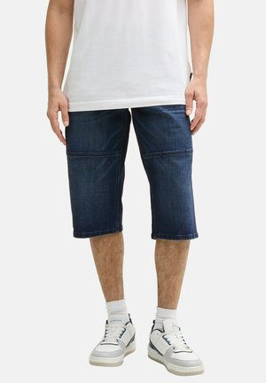 Jeans šortsevi, tamnoplave boje, do koljena, s vodoravnim šavovima. U kombinaciji s bijelom majicom i bijelim tenisicama s plavim detaljima.