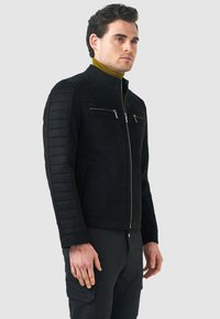 Giacca in suede nero con texture trapuntata sulle spalle, tasche con zip frontali e colletto rialzato. Indossata sopra un dolcevita senape.