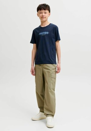 Dreng står iført en navy blå T-shirt med grafik og korte ærmer, khaki cargobukser og hvide sneakers mod en ensfarvet lys baggrund.