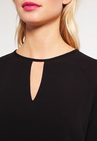 Top nero con scollo a chiave, colletto rotondo e texture liscia, con una leggera lucentezza e forma attillata intorno allo scollo.