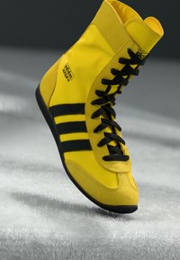 High-top sneakers made of yellow synthetic material, featuring black laces, three black stripes, and branding on the side. Textured rubber sole.