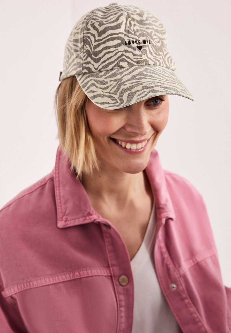 Femme souriante aux cheveux blonds courts portant une casquette beige et grise à motif zèbre et une veste rose boutonnée sur une chemise blanche.