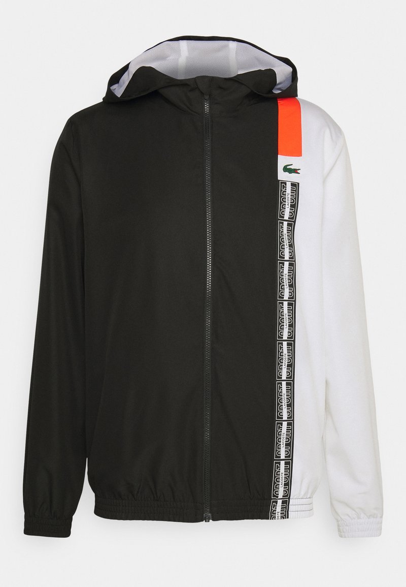 Giacca zip-up bianca e nera con cappuccio regolabile, caratterizzata da un accento arancione brillante e dettagli in nastro logo sui lati. Tessuto liscio e leggero.