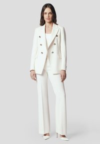 Biała marynarka double-breasted z złotymi guzikami, zestawiona z dopasowanymi białymi spodniami i szpilkami z wąskim noskiem. Gładki, wykończony materiał o strukturalnym designie.