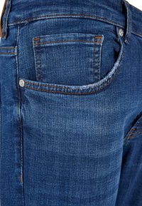 Jean en denim en gros plan montrant la poche arrière avec des coutures orange visibles et un rivet métallique sur un tissu bleu profond.
