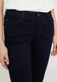 Jeans de mezclilla azul oscuro con corte ajustado, que incluyen bolsillos frontales, costuras en contraste y un cierre de botón en la cintura.