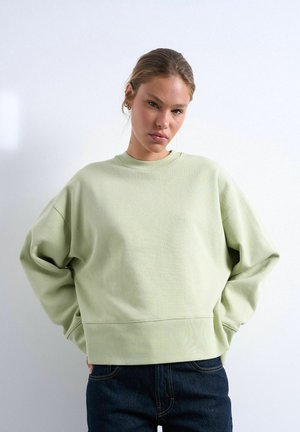 Ung kvinde iført en lysegrøn stor sweatshirt og mørke jeans står med hænderne på hoften mod en ensfarvet hvid baggrund.