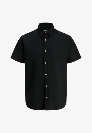 Garçon aux cheveux courts et bouclés portant une chemise noire à boutons à manches courtes, un pantalon gris ample et des baskets blanches, debout et souriant.