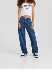 Top corto blanco con la impresión "POSITANO", combinado con pantalones cargo de denim azul sueltos que tienen bolsillos laterales y zapatillas negras.