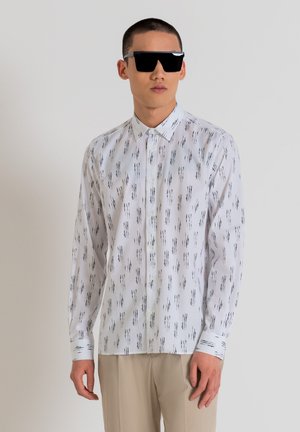 Camicia bianca con un motivo astratto nero, maniche lunghe e colletto. Il modello indossa occhiali da sole neri e pantaloni chiari.