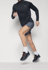 Svart långärmad idrottströja med kvart zip, grå shorts, vita strumpor och svarta löparskor med orange detaljer; i rörelse.