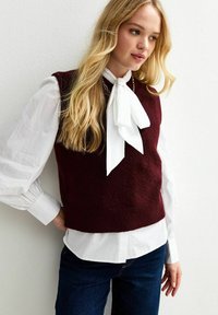 Burgundy stickad väst med en kortad design över en vit blus med en stor rosett. Strukturerad tyg och avslappnad passform.