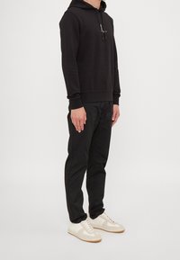Felpa nera con cappuccio e cordino, abbinata a pantaloni neri e sneakers bianche. L'outfit presenta un design semplice e minimalista.
