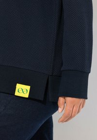 Mörkblå sweatshirt med textur och ribbade ärmslut, med en gul tagg med en grön oändlighetssymbol i nederkant.