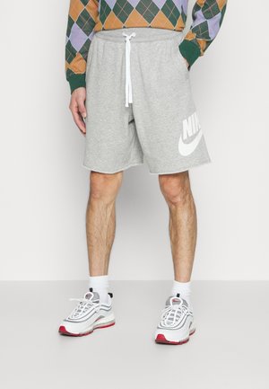 Persoon die grijze Nike-shorts met witte trekkoordjes draagt, een ruitjestrui, witte sokken en witte en rode sneakers, staand op een effen vloer.