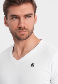 T-shirt bianca in cotone a V con un piccolo logo nero sul lato sinistro del petto. Il tessuto è liscio e ha una forma aderente.