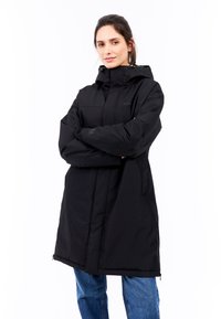 Sort isoleret frakke med høj krave, hætte og lynlås foran, med sidelommer og elastiske manchetter. Båret over blå jeans.