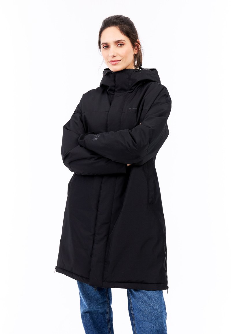 Sort isoleret frakke med høj krave, hætte og lynlås foran, med sidelommer og elastiske manchetter. Båret over blå jeans.