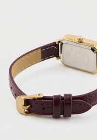 Auksinio atspalvio stačiakampis laikrodis su bordo odiniu dirželiu, puoštas rožine siūle ir poliruota metaline sagtimi. Dirželio paviršius turi reljefinį raštą.