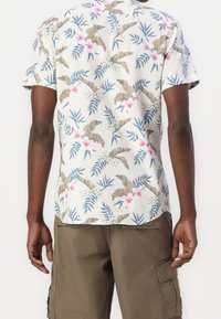 Camisa de manga corta en tela ligera con un estampado floral tropical que incluye hojas verdes, flores rosas y detalles marrones. Se llevan puestos pantalones cortos cargo caqui.