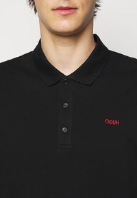 Mladý člověk v černém polo tričku na knoflíky s červeným vyšitým logem na levé hrudi, které nese nápis "HUGO" obráceně.