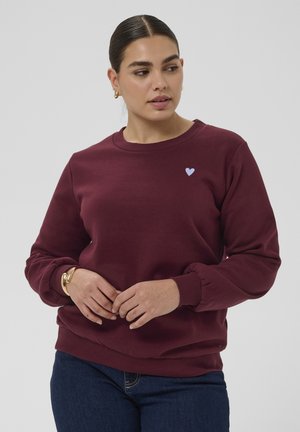 Donna con capelli scuri raccolti in uno chignon, indossa una felpa color vinaccia con un piccolo cuore azzurro chiaro sul petto e jeans blu scuro, con le mani intrecciate.