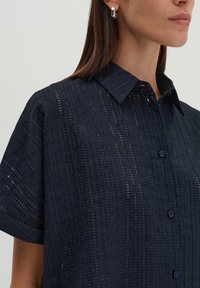 Camicia blu navy con un motivo testurizzato. Presenta un colletto a punta e maniche corte. Include bottoni blu lungo la pattina.