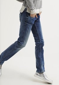 Vaqueros de mezclilla azul oscuro con corte ajustado y un acabado desgastado. Combinados con una blusa de rayas y zapatos plateados de tipo slip-on.