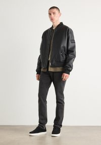 Zwarte faux leren bomberjack met ribgebreide manchetten, rits aan de voorkant en zijzakken. Draagt over een beige top, gecombineerd met donkere jeans en zwarte sneakers.