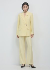 Completo giallo su misura con giacca a un bottone, due tasche frontali e pantaloni a gamba larga coordinati. Completato con tacchi a punta neri.