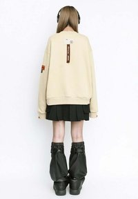 Sudadera beige de gran tamaño con etiqueta, falda negra plisada y botas negras brillantes y ajustadas. Cabello cubierto por auriculares, visto desde atrás.