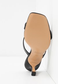Sandale noire à talon haut avec bout carré et semelle beige, inclinée contre un coin blanc, affichant « Vero Cuoio Made in Italy » et taille 35.