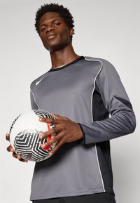 Grå langermet treningsgenser med svarte mesh-detaljer og hvite kanter, holdt av en person med en mønstrete fotball med røde detaljer.