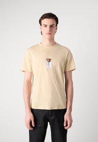 Beige bomulds t-shirt med et grafisk print af en hund og ordet "Hugo" i midten, parret med mørk denim jeans.