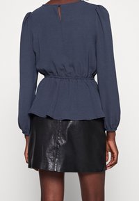 Femme portant un chemisier bleu marine à manches longues avec une taille froncée et une mini-jupe en cuir noir, vue de dos sur un fond uni.