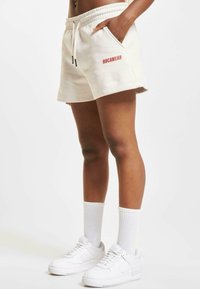 Beige cotton shorts with elastic waistband, side pockets, and red "ROCAWEAR" logo. Paired with white sneakers and ribbed white socks.